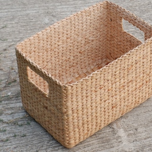 Puede incluir: Una cesta de almacenamiento tejida con dos asas recortadas. La cesta está hecha de un material natural de color marrón claro y tiene una forma rectangular.