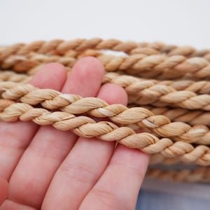Puede incluir: Primer plano de una mano sosteniendo un manojo de cuerda de fibra natural. La cuerda es de color marrón claro y tiene una textura trenzada y retorcida.