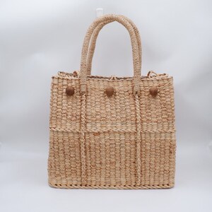 K&ouml;nnte beinhalten: Eine geflochtene Strohtasche mit braunen Holz-Knopf-Akzenten. Die Tasche hat eine rechteckige Form mit zwei oberen Griffen.