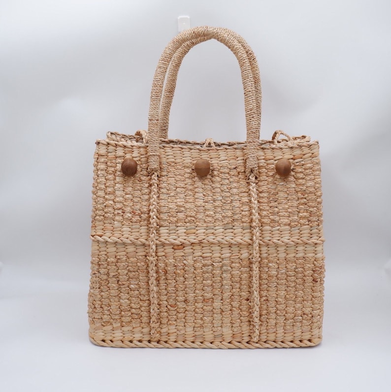 K&ouml;nnte beinhalten: Eine geflochtene Strohtasche mit braunen Holz-Knopf-Akzenten. Die Tasche hat eine rechteckige Form mit zwei oberen Griffen.