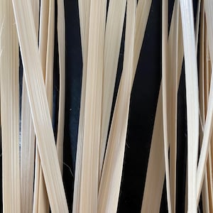 May include: A close-up of a bundle of natural, light brown, woven, flat, and flexible strips of material. The strips are arranged vertically and are slightly overlapping.