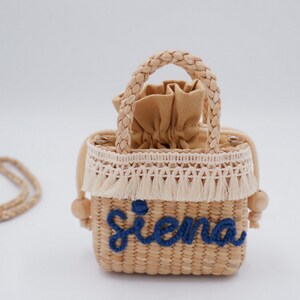 Op de afbeelding: Een kleine, geweven strooien handtas met een gevlochten handvat en een afneembare schouderriem. De tas heeft een beige trekkoordsluiting, een crèmekleurige franje en de naam "Siena" geborduurd in blauw. Houten kralen accentueren de zijkanten.