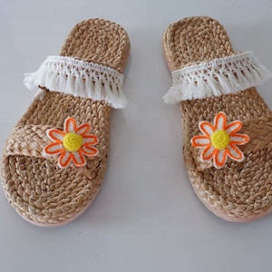 May include: A pair of brown woven slide sandals with white fringe and orange flower embellishments.