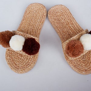May include: A pair of brown woven slippers with three pom-poms on each slipper. The pom-poms are brown, white, and dark brown.