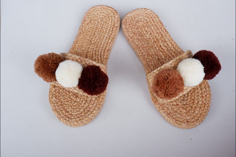 May include: A pair of brown woven slippers with three pom-poms on each slipper. The pom-poms are brown, white, and dark brown.