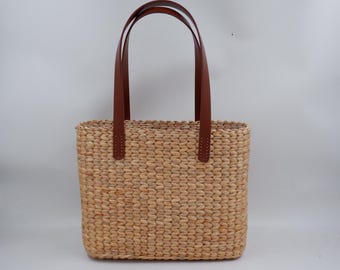 May include: A woven straw tote bag with brown leather handles. The bag is a light brown colour and has a rectangular shape.