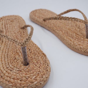 Op de afbeelding: Een paar geweven strooien slippers met een eenvoudig ontwerp. De sandalen zijn lichtbruin van kleur en hebben een platte zool.