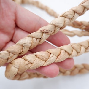 May include: Close-up of a natural, beige, braided rope made from woven straw. The rope is thick and has a natural, rustic look.