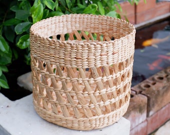 May include: A natural woven basket with a round shape and a lattice pattern. The basket is made of light brown, natural materials and has a rustic, handmade look.