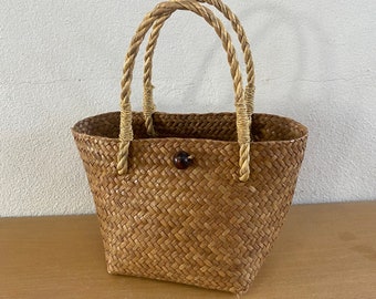 May include: A woven basket bag with a trapezoidal shape. The bag is made of light brown woven material with two braided handles. A dark brown button secures the bag's closure. The bag sits on a light brown wooden surface.