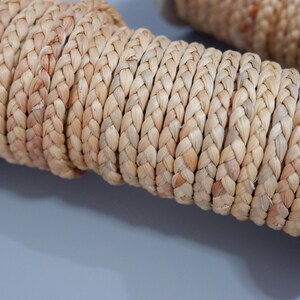 Könnte beinhalten: Nahaufnahme von zwei Spulen aus natürlichem, gewebtem Bastband. Das Band ist eng geflochten und zeigt eine hellbraune Farbe mit subtilen Variationen. Die Spulen sind teilweise sichtbar, was auf einen Bastel- oder Dekorationsartikel hindeutet.