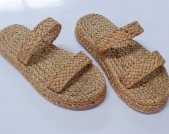 May include: A pair of tan sandals crafted from woven natural fibers. Each sandal features a thick, braided sole and two wide, braided straps across the top. The construction is uniform throughout, showcasing a consistent braided texture in a light brown, earthy tone.