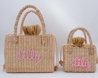 May include: Two straw tote bags with pink embroidered names "Lilly". The larger bag has a double handle and the smaller bag has a single handle.