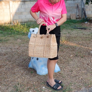 K&ouml;nnte beinhalten: Eine geflochtene Strohtasche mit braunen Ledergriffen. Die Tasche wird von einer Person gehalten, die ein rosa Poloshirt und einen schwarzen Rock tr&auml;gt.