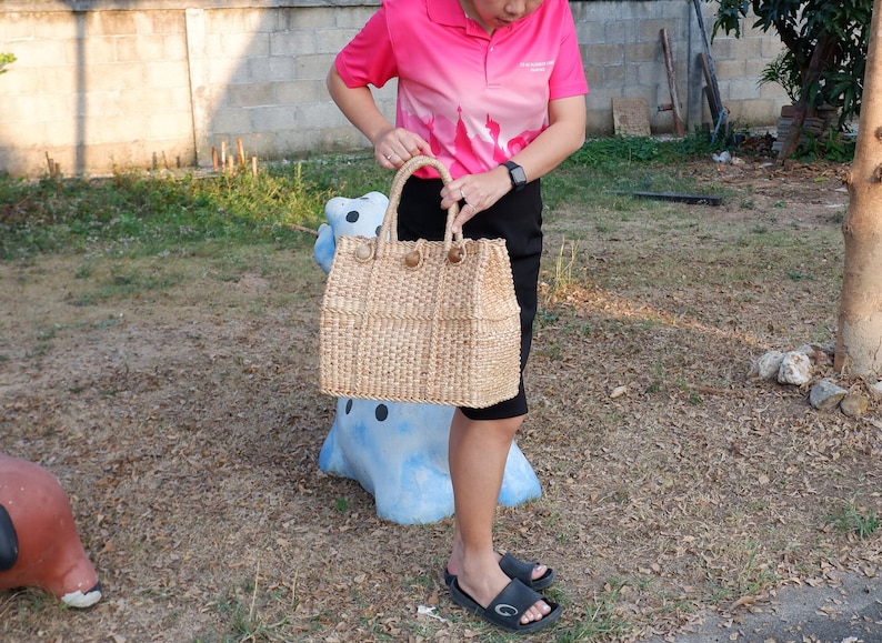K&ouml;nnte beinhalten: Eine geflochtene Strohtasche mit braunen Ledergriffen. Die Tasche wird von einer Person gehalten, die ein rosa Poloshirt und einen schwarzen Rock tr&auml;gt.