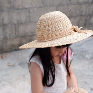Pode incluir: Uma menina pequena usando um vestido branco e um chapéu de palha grande e tecido com um detalhe de flor rosa. O chapéu tem uma aba larga e é feito de materiais naturais.