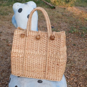 K&ouml;nnte beinhalten: Eine geflochtene Strohtasche mit braunen Holzgriffen und braunen Holzkn&ouml;pfen. Die Tasche liegt auf einer blau-wei&szlig; gepunkteten Oberfl&auml;che.