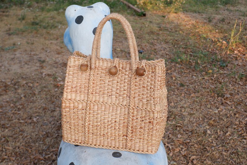 K&ouml;nnte beinhalten: Eine geflochtene Strohtasche mit braunen Holzgriffen und braunen Holzkn&ouml;pfen. Die Tasche liegt auf einer blau-wei&szlig; gepunkteten Oberfl&auml;che.