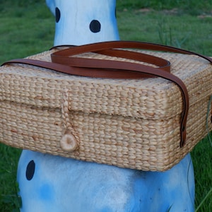 May include: A woven straw tote bag with brown leather straps. The bag is sitting on a blue and white surface.