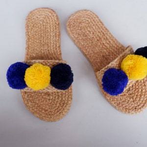 May include: A pair of tan jute slides with blue and yellow pom poms. The slides are made of woven jute and have a simple design. The pom poms are attached to the front of the slides and are a fun and colorful addition.