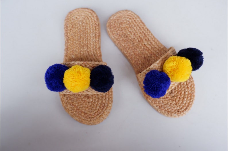 May include: A pair of tan jute slides with blue and yellow pom poms. The slides are made of woven jute and have a simple design. The pom poms are attached to the front of the slides and are a fun and colorful addition.