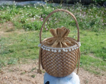 May include: A woven straw bag with a brown fabric lining and white pom-poms around the top. The bag has a brown woven handle.