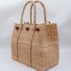 K&ouml;nnte beinhalten: Eine geflochtene Strohtasche mit zwei Henkeln und Holz-Knopf-Akzenten. Die Tasche ist aus nat&uuml;rlichem, hellbraunem Material.