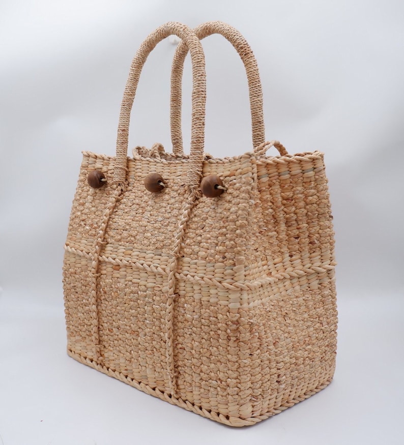 K&ouml;nnte beinhalten: Eine geflochtene Strohtasche mit zwei Henkeln und Holz-Knopf-Akzenten. Die Tasche ist aus nat&uuml;rlichem, hellbraunem Material.