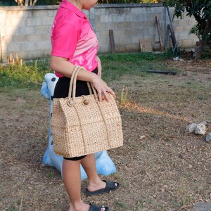 K&ouml;nnte beinhalten: Eine Frau h&auml;lt eine gro&szlig;e, geflochtene Strohtasche mit braunen Ledergriffen und Kn&ouml;pfen. Die Tasche hat eine rechteckige Form und ist aus einem nat&uuml;rlichen, hellbraunen Material gefertigt.