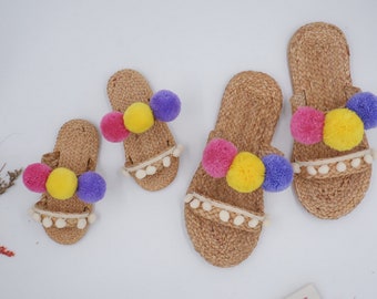 May include: Three pairs of straw sandals with colorful pom-poms. The sandals are in different sizes, with the smallest pair having purple, yellow, and pink pom-poms.