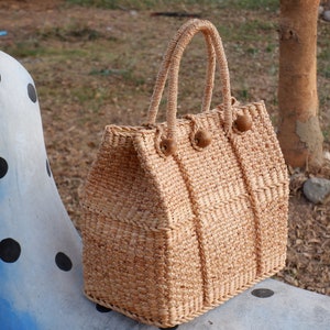 K&ouml;nnte beinhalten: Eine geflochtene Strohtasche mit Holz-Knopfverschl&uuml;ssen. Die Tasche ist hellbraun und steht auf einer blau-wei&szlig;en Oberfl&auml;che mit Punkten.