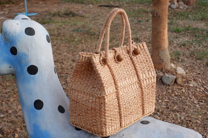 K&ouml;nnte beinhalten: Eine geflochtene Strohtasche mit Holz-Knopfverschl&uuml;ssen. Die Tasche ist hellbraun und steht auf einer blau-wei&szlig;en Oberfl&auml;che mit Punkten.