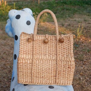 K&ouml;nnte beinhalten: Eine geflochtene Strohtasche mit braunen Holzgriffen. Die Tasche hat eine rechteckige Form und ist aus nat&uuml;rlichem Strohmaterial gefertigt.