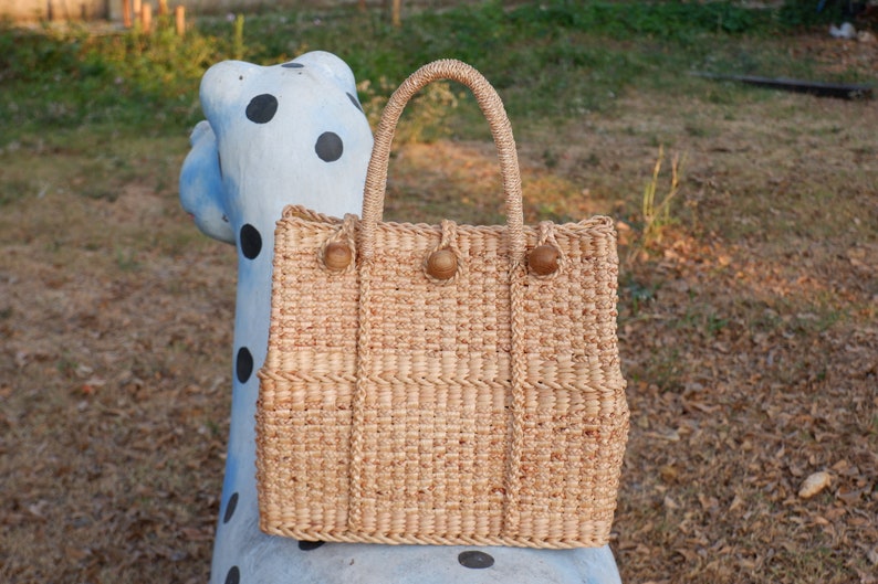 K&ouml;nnte beinhalten: Eine geflochtene Strohtasche mit braunen Holzgriffen. Die Tasche hat eine rechteckige Form und ist aus nat&uuml;rlichem Strohmaterial gefertigt.