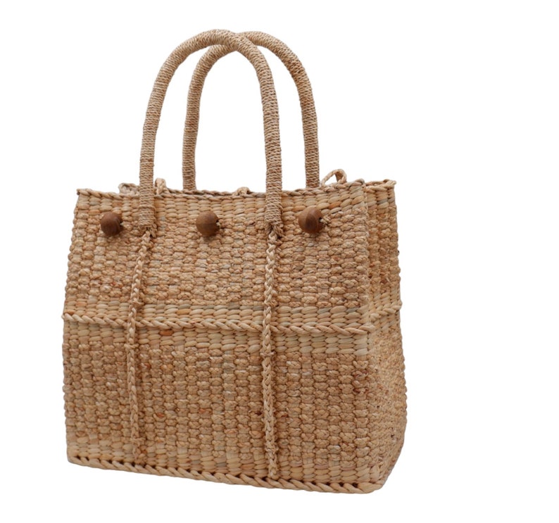 K&ouml;nnte beinhalten: Eine geflochtene Strohtasche mit braunen Holz-Knopf-Akzenten. Die Tasche hat zwei Henkel und eine breite &Ouml;ffnung.