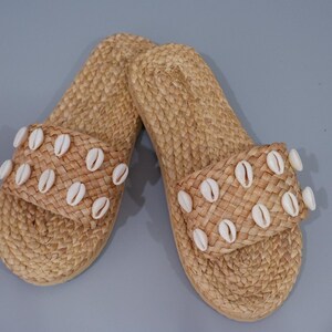 Op de afbeelding: Een paar bruine geweven sandalen met witte schelpversieringen. De sandalen hebben een eenvoudig ontwerp en zijn perfect voor de zomer.