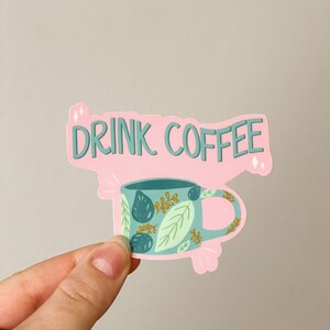 May include: Pink sticker with the text "DRINK COFFEE" and a blue coffee mug with a green and gold leaf pattern.