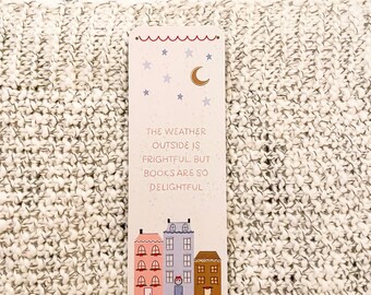May include: A rectangular bookmark with a whimsical design. The top features a red scalloped border with stars and a crescent moon. The text reads, "The weather outside is frightful, but books are so delightful." Below are illustrated houses.