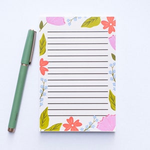 May include: A white notepad with a floral border. The border features pink, red, and green flowers and leaves. The notepad has lined paper.