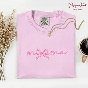 May include: A light pink Comfort Colors t-shirt with the word "mama" in a cursive font and a bow design. A gold necklace with a star pendant rests on the shirt. The words "Designer Park Special Designs" are in the upper right corner.
