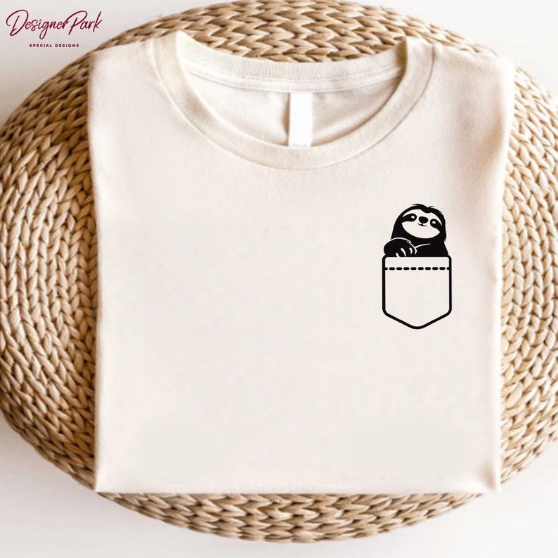 Puede incluir: Camiseta color crema con un gr&aacute;fico negro de un perezoso asomando por un bolsillo. El perezoso tiene una cara sonriente y est&aacute; posicionado en un dise&ntilde;o de bolsillo. La camiseta tiene cuello redondo y mangas cortas.