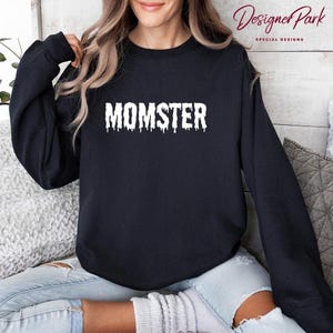 A woman wearing a black sweatshirt with the word "MOMSTER" printed on it, sitting on a couch.