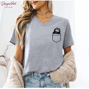 Puede incluir: Camiseta gris con cuello en V con un dise&ntilde;o de perezoso negro en un bolsillo. La camiseta est&aacute; hecha de un material suave y tiene mangas cortas. La camiseta se combina con unos vaqueros azules.