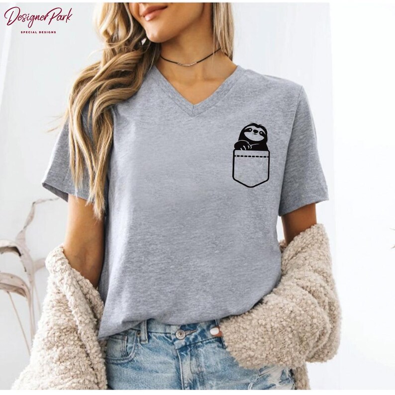 Puede incluir: Camiseta gris con cuello en V con un dise&ntilde;o de perezoso negro en un bolsillo. La camiseta est&aacute; hecha de un material suave y tiene mangas cortas. La camiseta se combina con unos vaqueros azules.