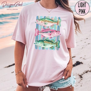 A woman wearing a pink t-shirt with a graphic of three colorful fish printed on it, standing on a beach.