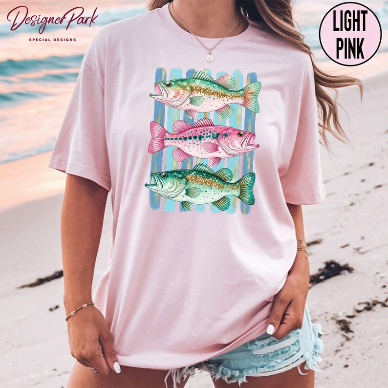 A woman wearing a pink t-shirt with a graphic of three colorful fish printed on it, standing on a beach.
