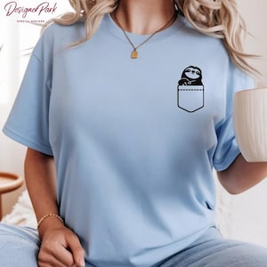 Puede incluir: Camiseta azul claro con un dise&ntilde;o de perezoso negro en un bolsillo. La camiseta tiene mangas cortas y cuello redondo. La marca "Designer Park" es visible en la esquina superior izquierda.