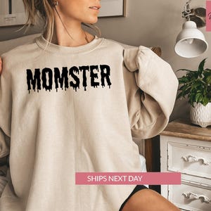 A woman wearing a beige sweatshirt with the word "MONSTER" printed on it, sitting on a bed next to a potted plant.