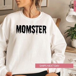 A woman wearing a white sweatshirt with the word "MOMSTER" printed on it, sitting in a room with a potted plant and a lamp.