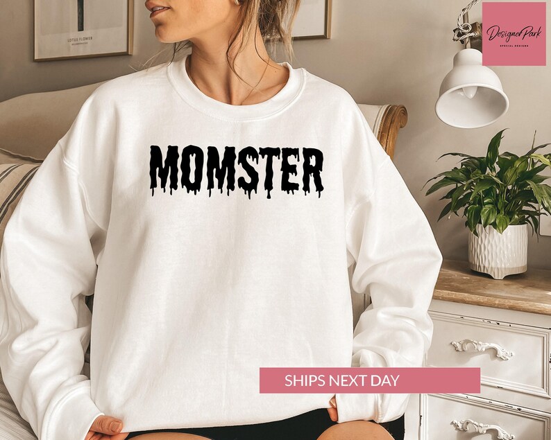 A woman wearing a white sweatshirt with the word "MOMSTER" printed on it, sitting in a room with a potted plant and a lamp.